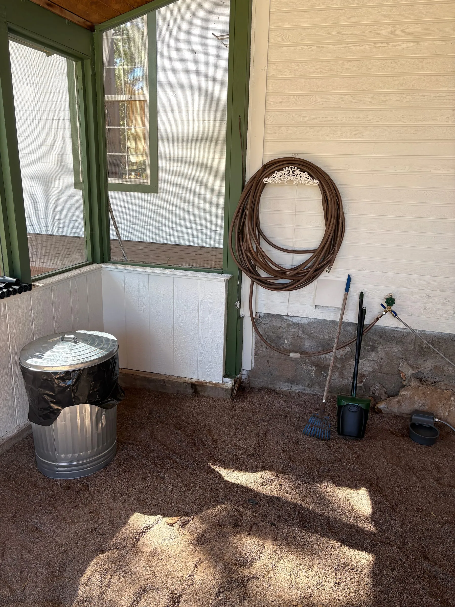 Inside the dog run — a lidded metal trash can with a liner in it, a coiled hose on the wall, and a rake and dustpan standing in the corner. The floor is decomposed granite.