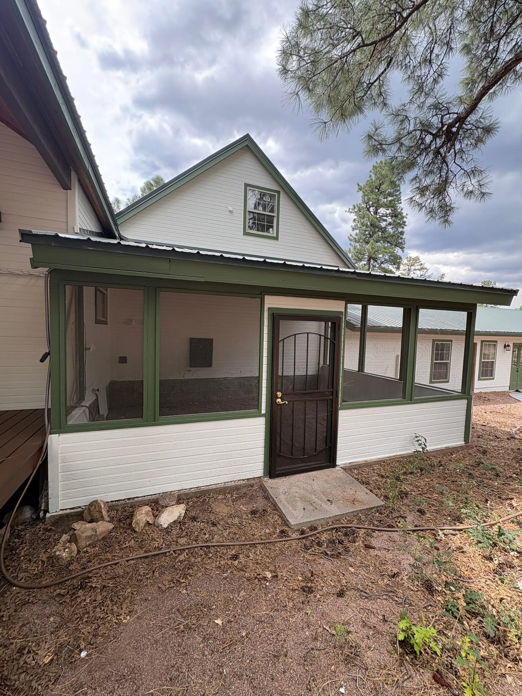 The dog run on the side of the lodge — a long screened enclosure under the roofline, framed in green-painted posts, with a screen door in the middle of it and pines behind.