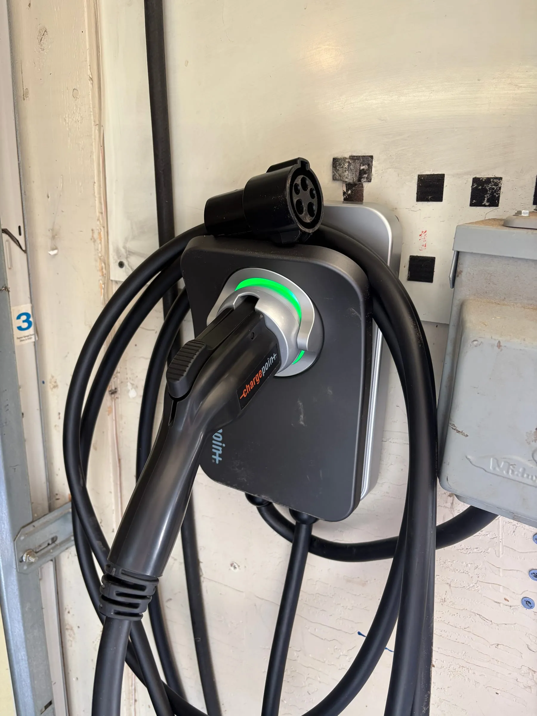 A ChargePoint Level 2 EV charger mounted on the garage wall between the two doors, its cable coiled beside it and the J1772 connector docked. A green ring on the unit shows it is live.