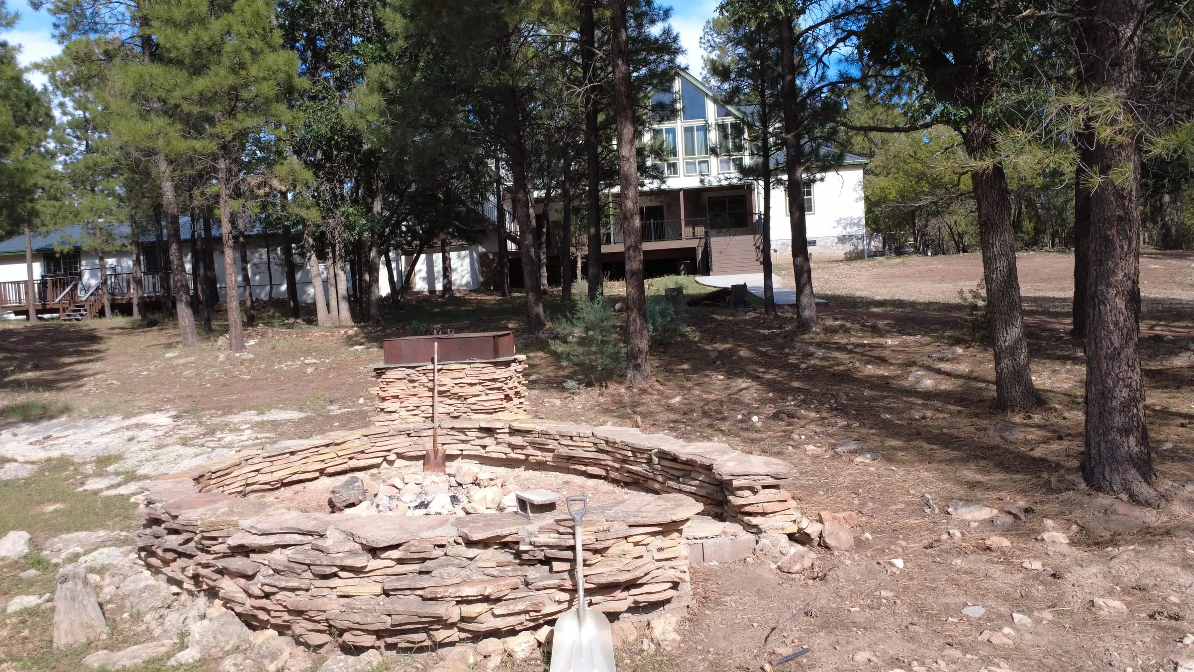 The firepit in the back yard — a wide, dry-stacked flagstone ring with a raised seating wall around it and a steel grate over one side. The lodge and the guest house stand in the pines behind it.
