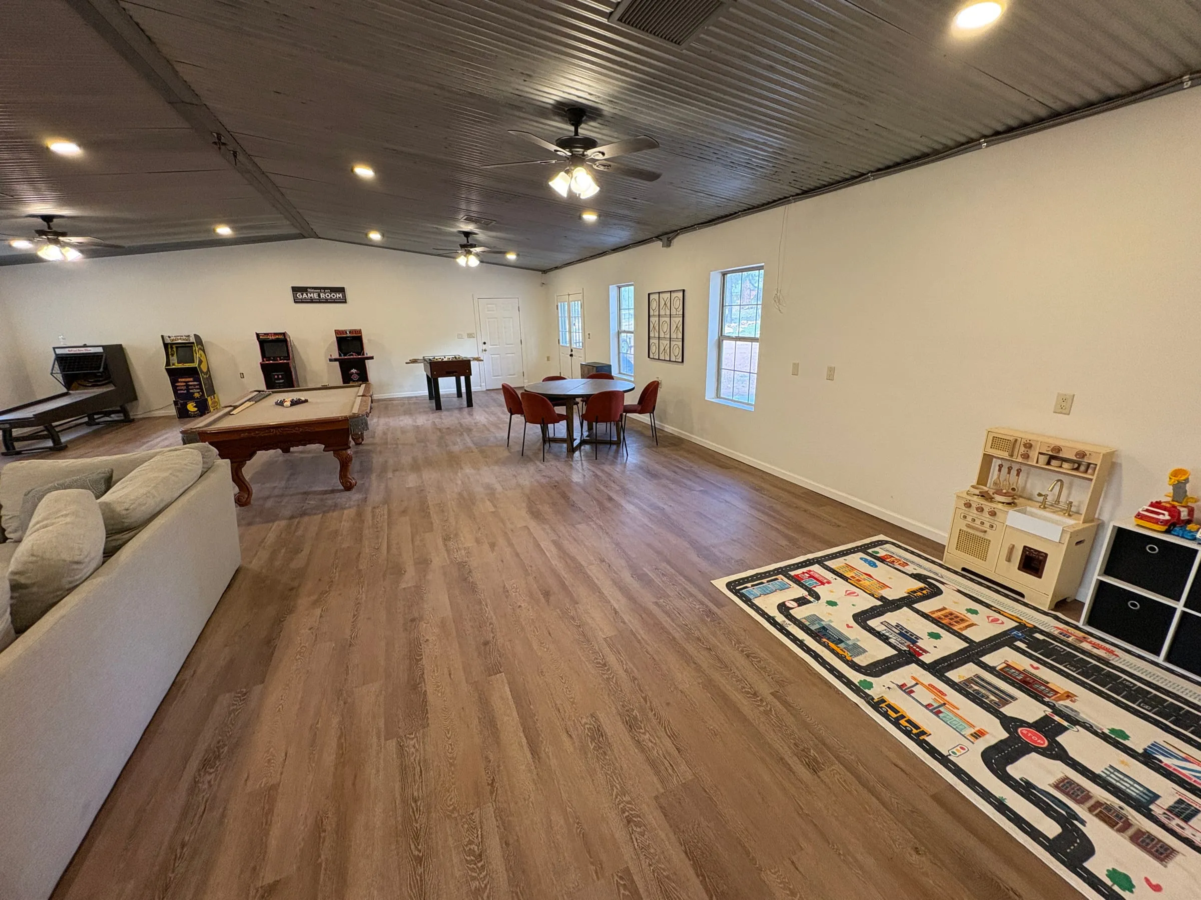 The game room from the far end — the billiards table and arcade cabinets on the left, a round card table with six chairs in the middle, and a children’s play area with a play kitchen and a road-map rug on the right.