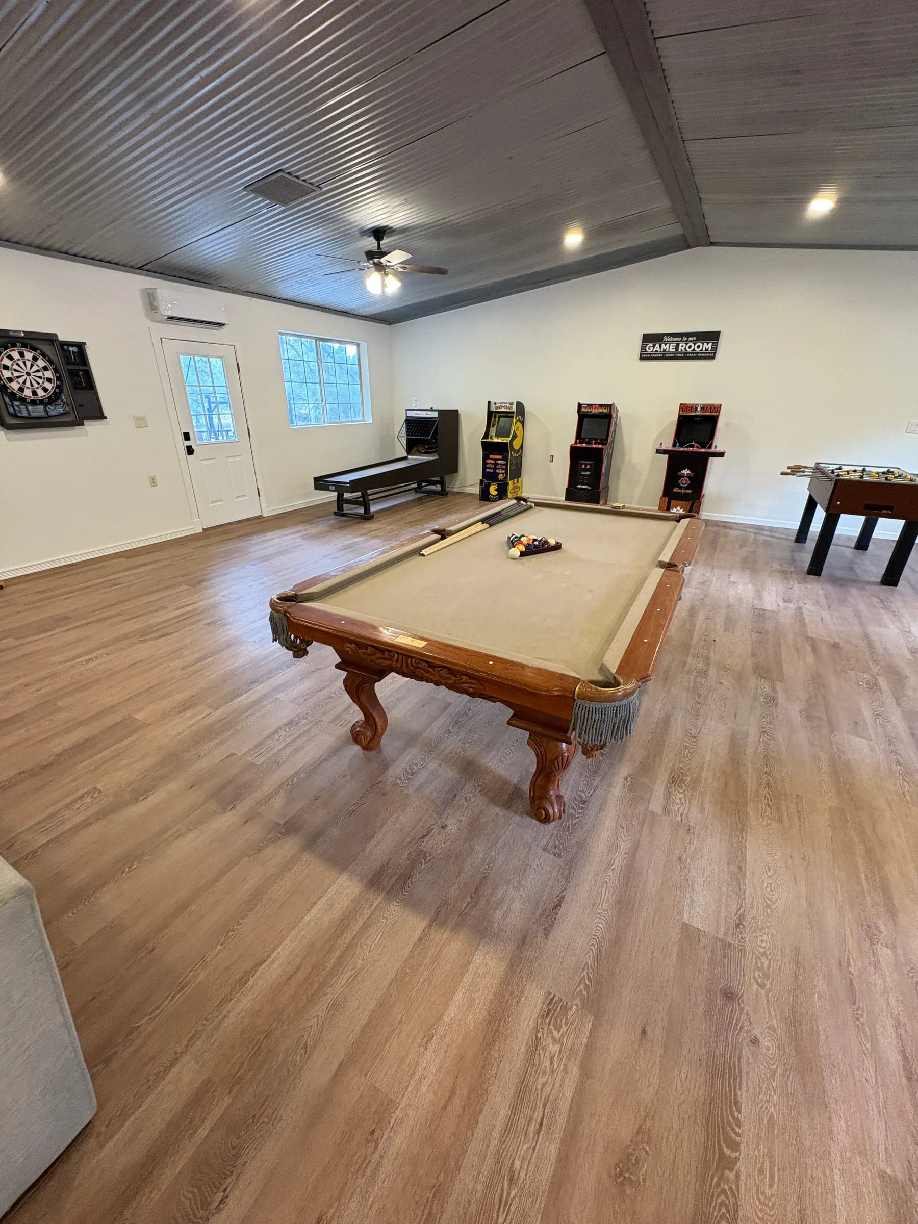A full-size billiards table with carved legs, racked and ready, with cues along the rail and the arcade cabinets and dart board behind it.