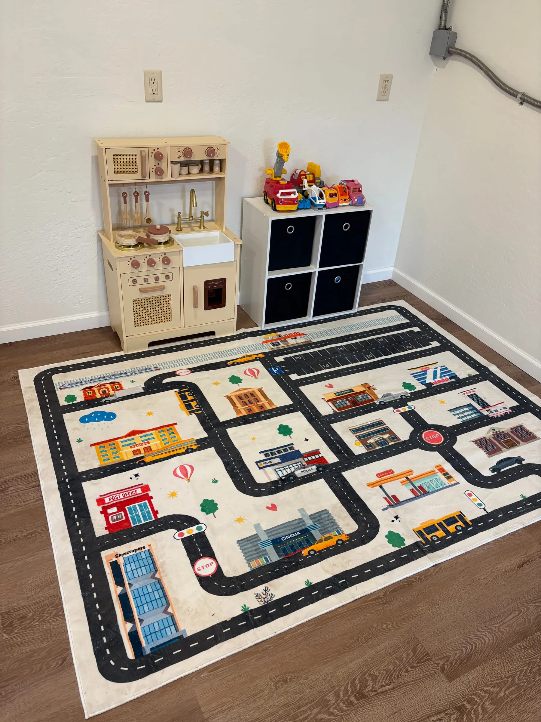 The children’s play area in the game room — a wooden play kitchen, a bin of toy cars and trucks, a four-cube storage shelf, and a large road-map rug.