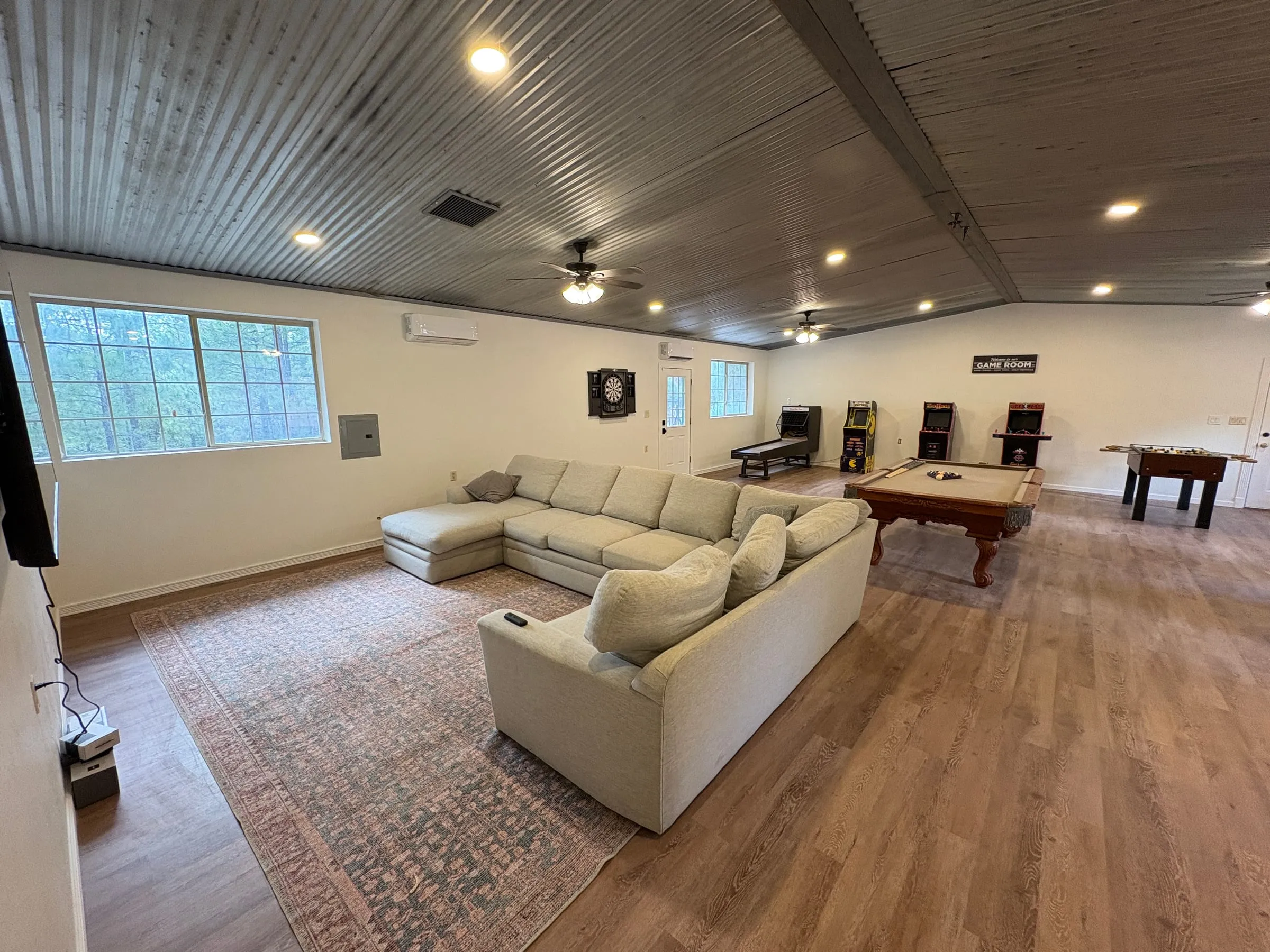 The game room — a long room under a corrugated metal ceiling, with a large sectional sofa and a wall-mounted TV at one end and a billiards table, three arcade cabinets, a skee-ball lane, a foosball table, and an electronic dart board at the other.
