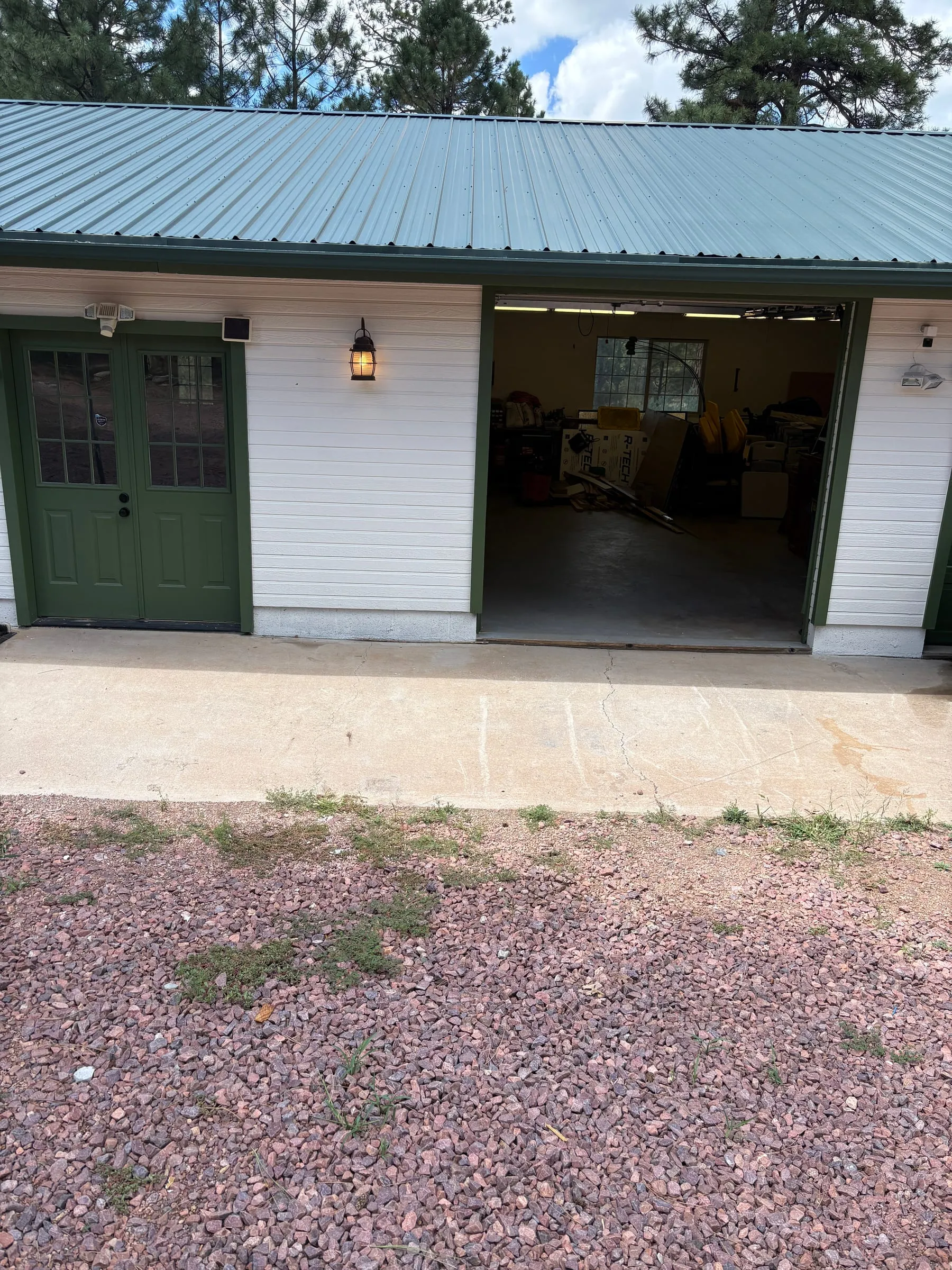 The attached garage from the gravel drive, with two doors side by side. One stands open. The charger is on the wall between them, inside.