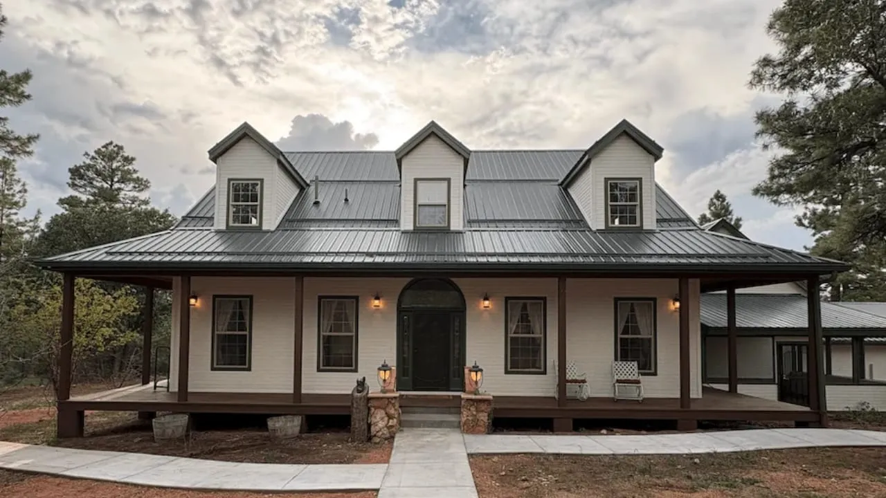 The front of Spoonbill Lodge at dusk — a two-story home with three dormers, a standing-seam metal roof, and a covered porch that wraps the front. Lanterns are lit beside the arched front door, and pines stand behind the house.