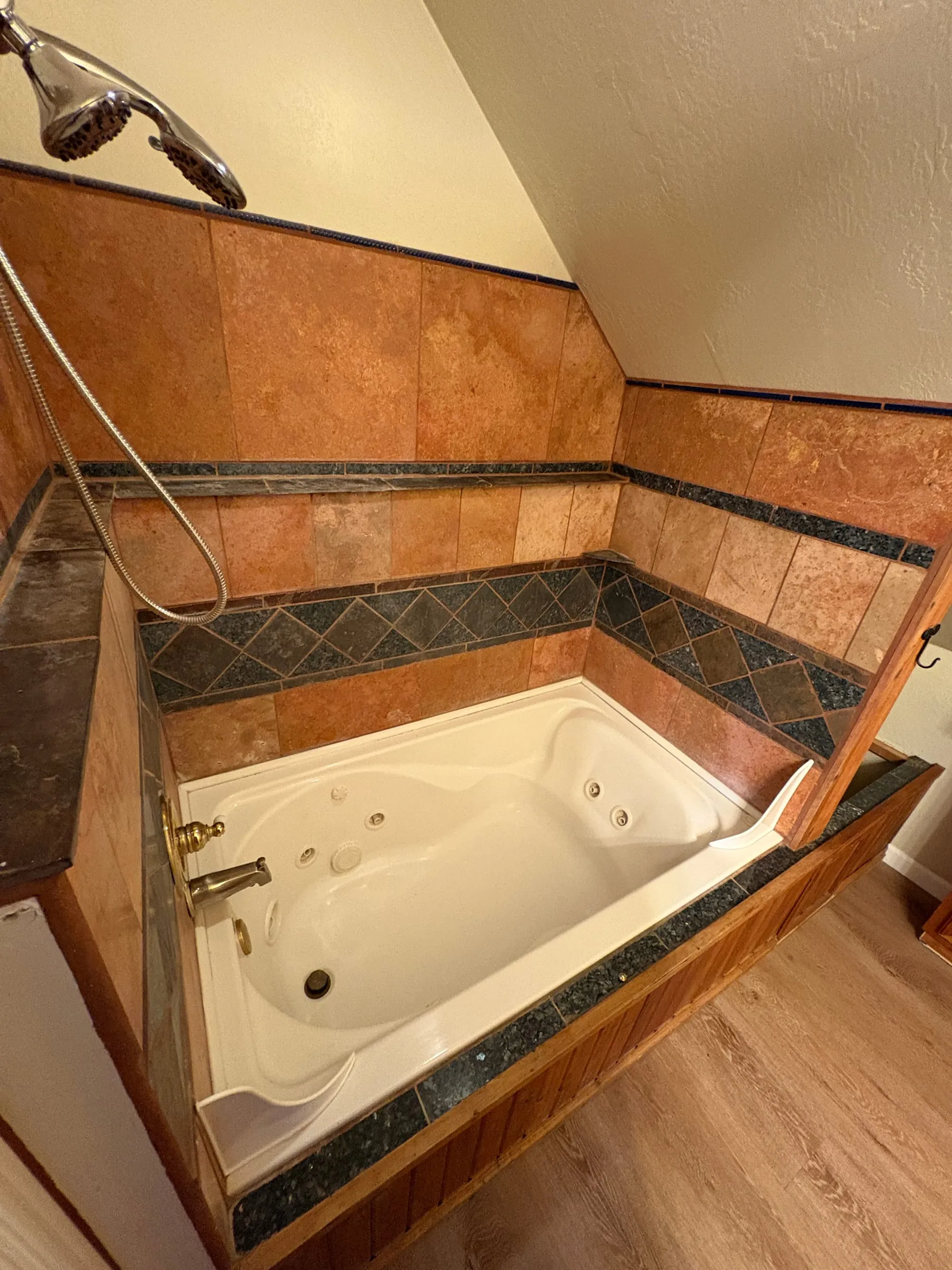 The bathroom in Nora’s studio — a jetted corner tub set into a tiled surround, with a hand-held shower head and slate accent tile.