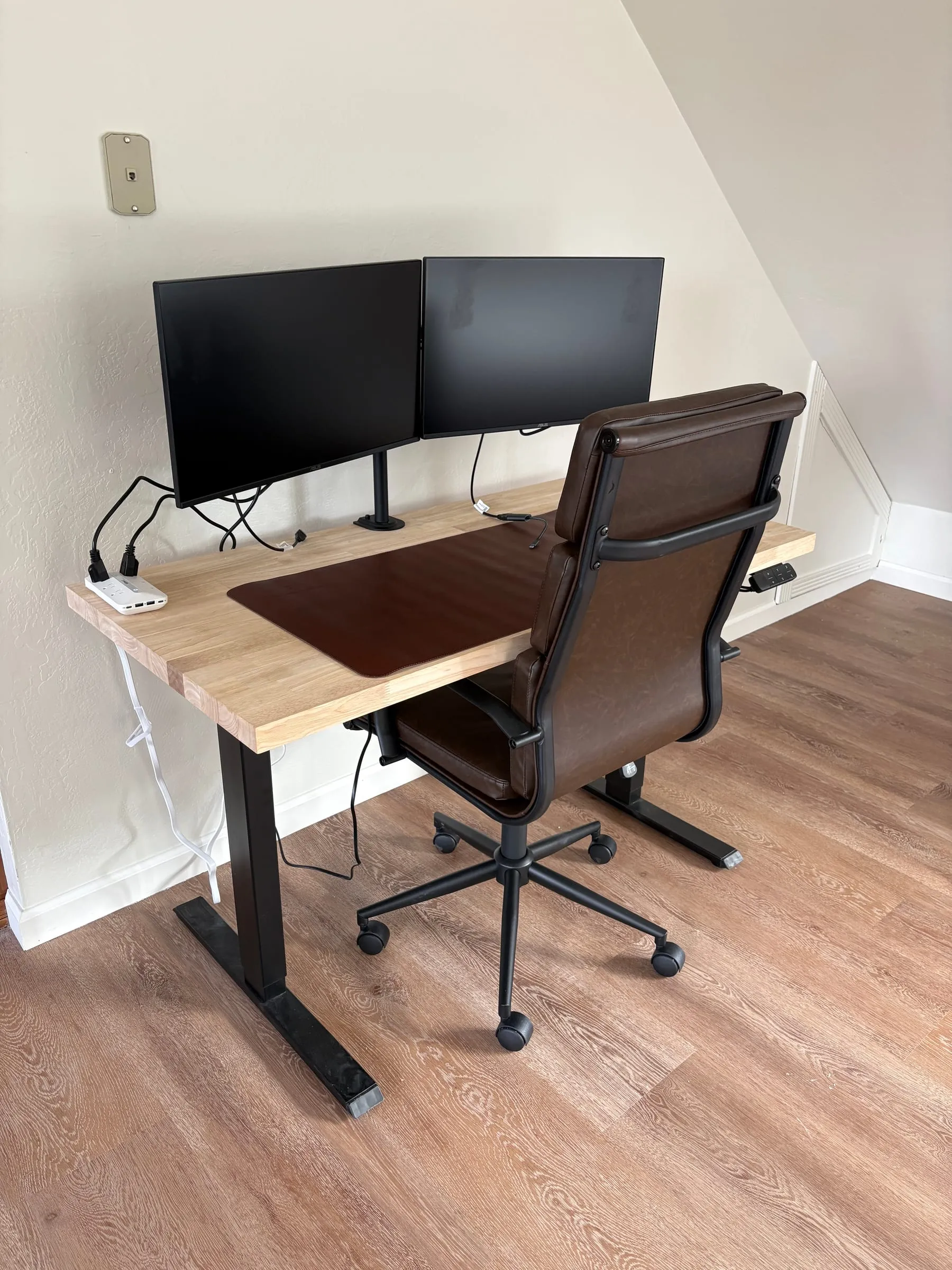 The work station in Nora’s studio — a height-adjustable standing desk with two monitors, a high-back leather chair, and a power strip on the desk. HDMI cables are there to connect your own computer.