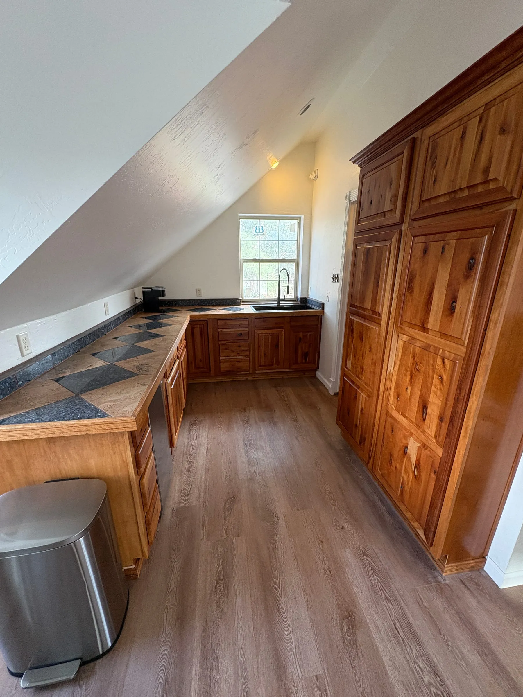 The kitchenette in Nora’s studio — knotty pine cabinets over a tiled counter, with a cooktop under wooden covers, a pantry cupboard, and a bin beside it.