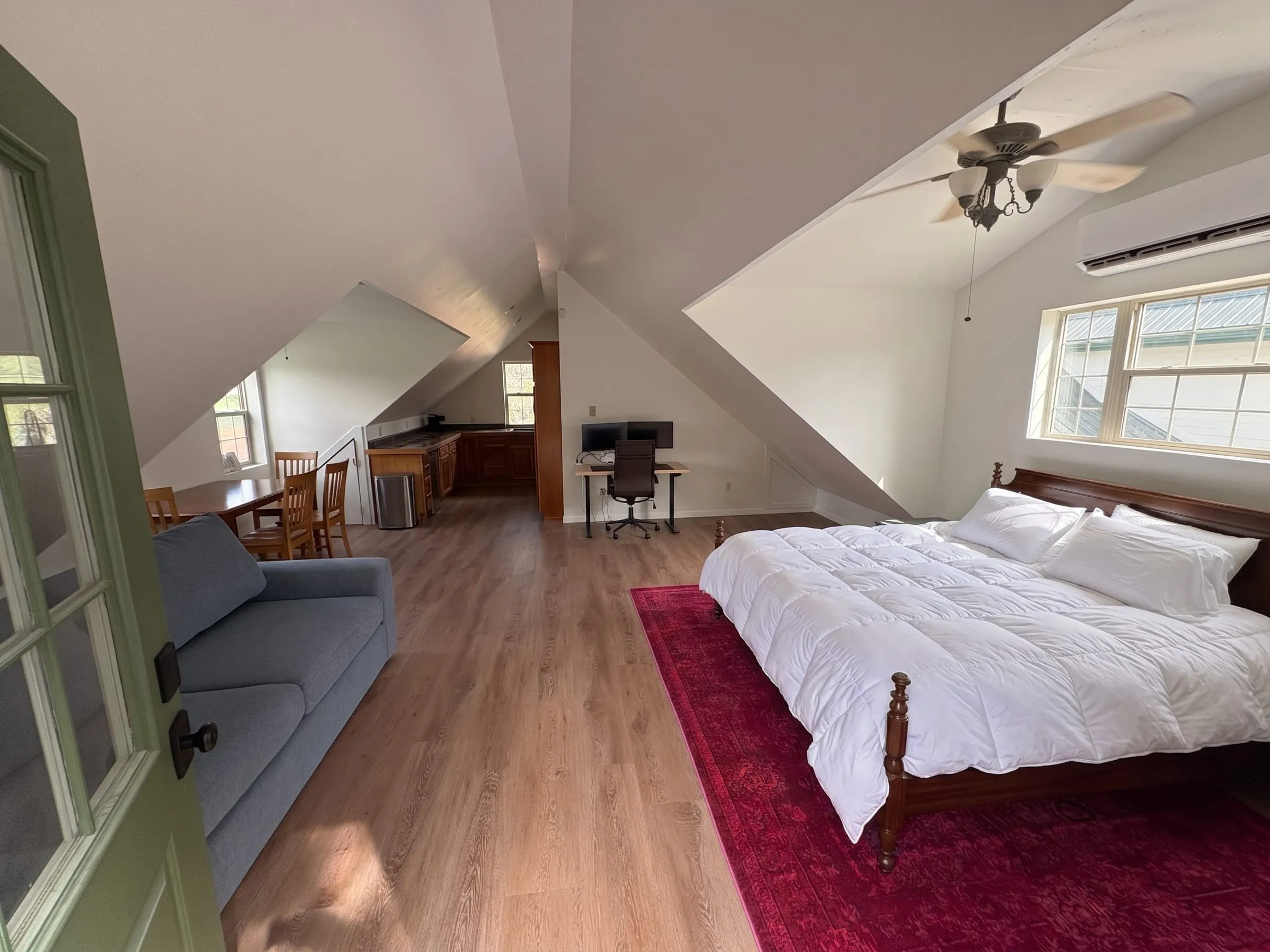 Nora’s studio seen from the door — one open room under a vaulted ceiling. A king bed sits on a red rug on the right, under a window and a mini-split unit. A sofa, a dining table, a kitchenette, and a standing desk run down the left side.