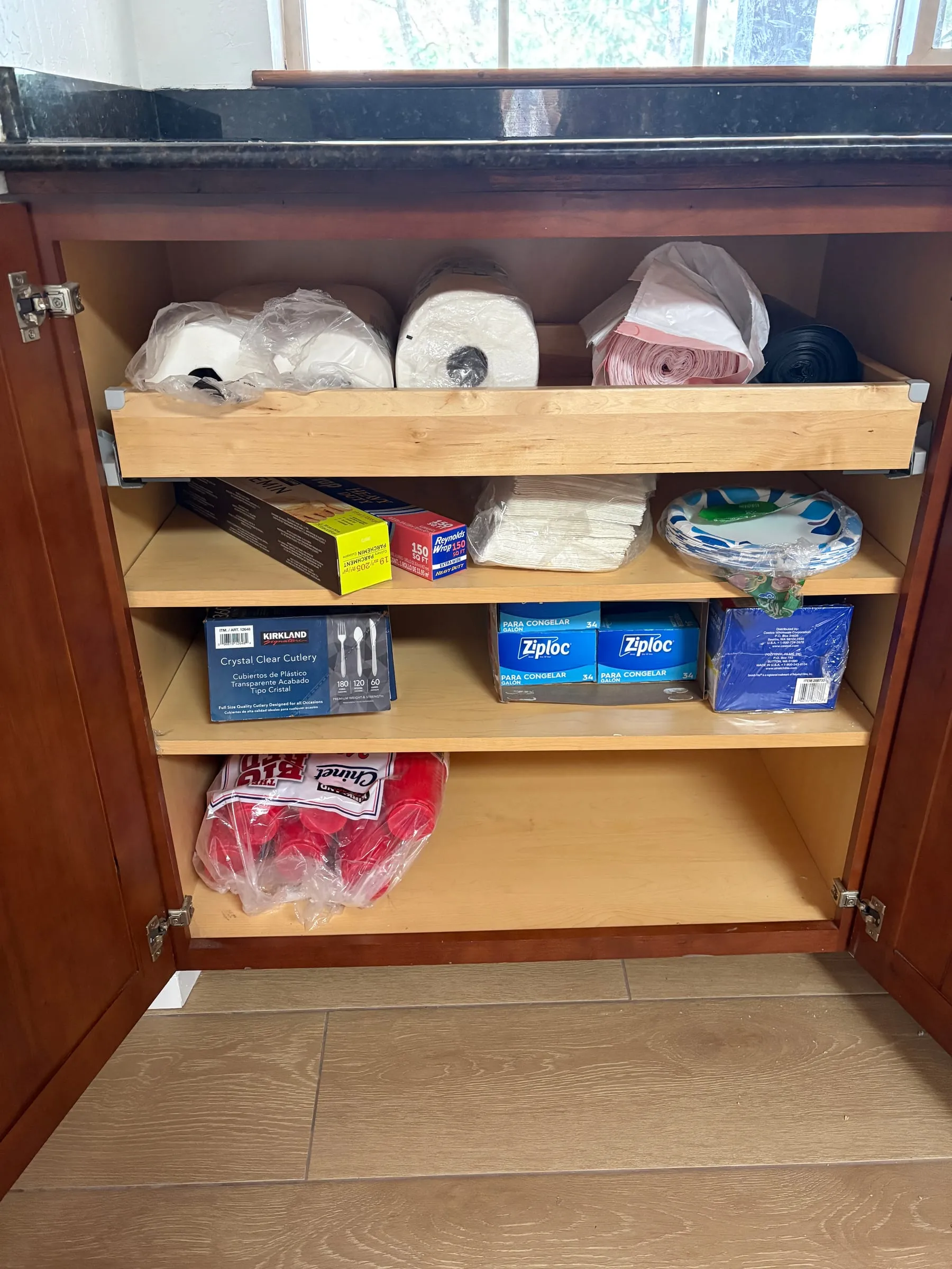 An open kitchen cabinet stocked with paper towels, toilet paper, napkins, paper plates, plastic cups, plastic cutlery, foil, parchment, zip bags, and rolls of trash bags.