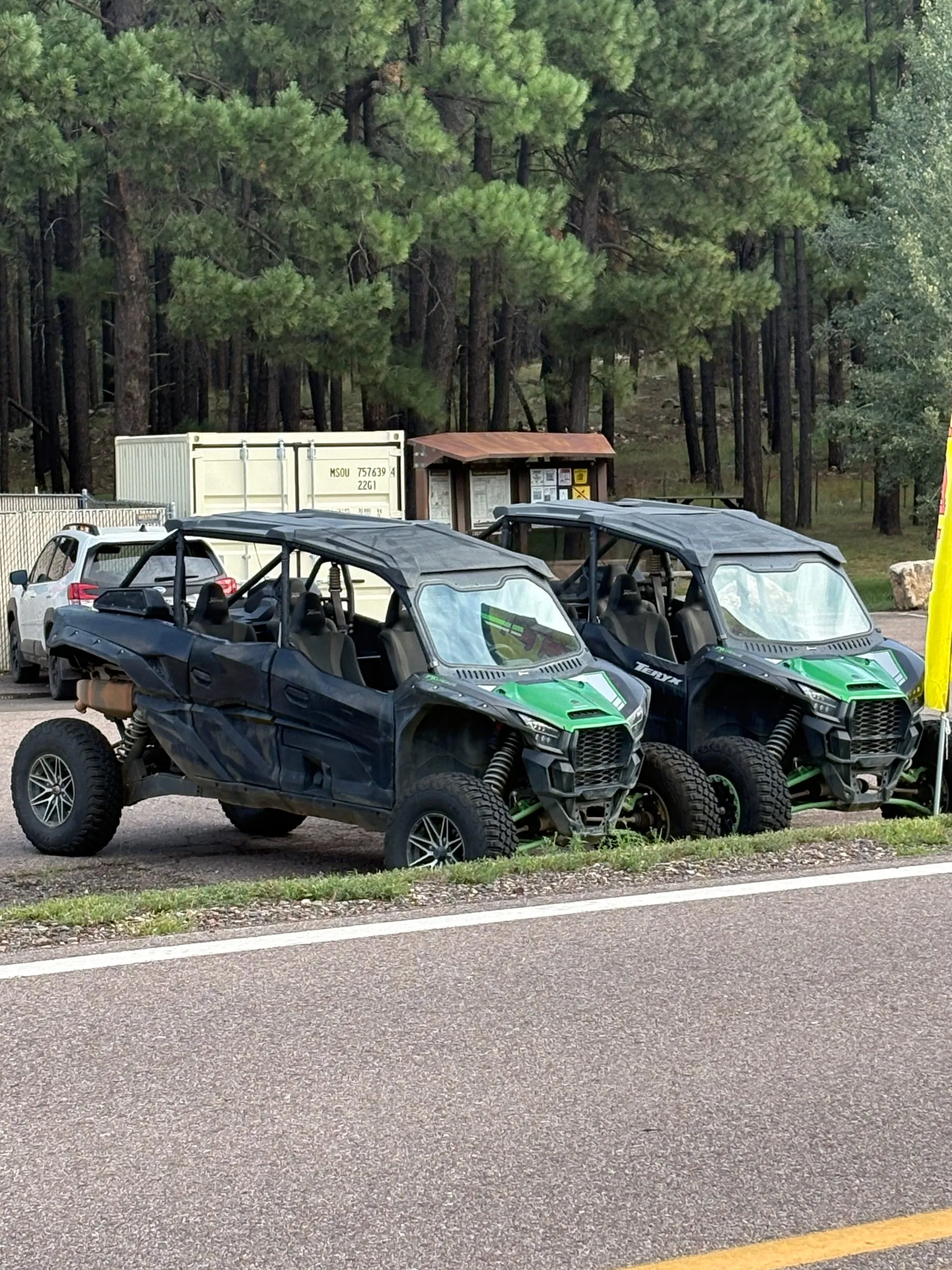 Two green four-seat side-by-sides parked at the roadside rental lot near Clints Well, both with roofs and windshields on them.