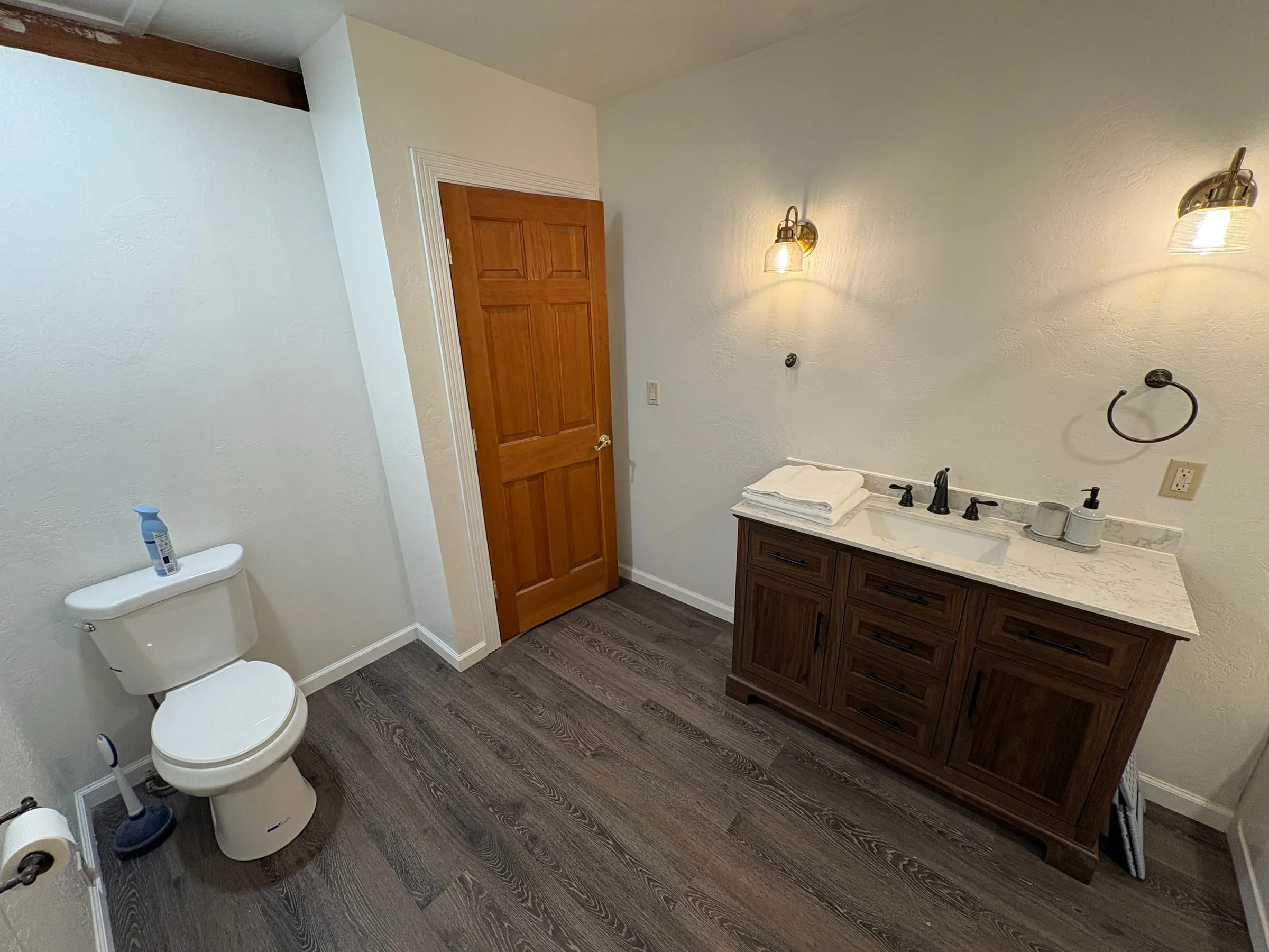 The theater bathroom from the other side — a wide vanity with a marble top, folded towels, and two brass sconces, with the toilet on the opposite wall.