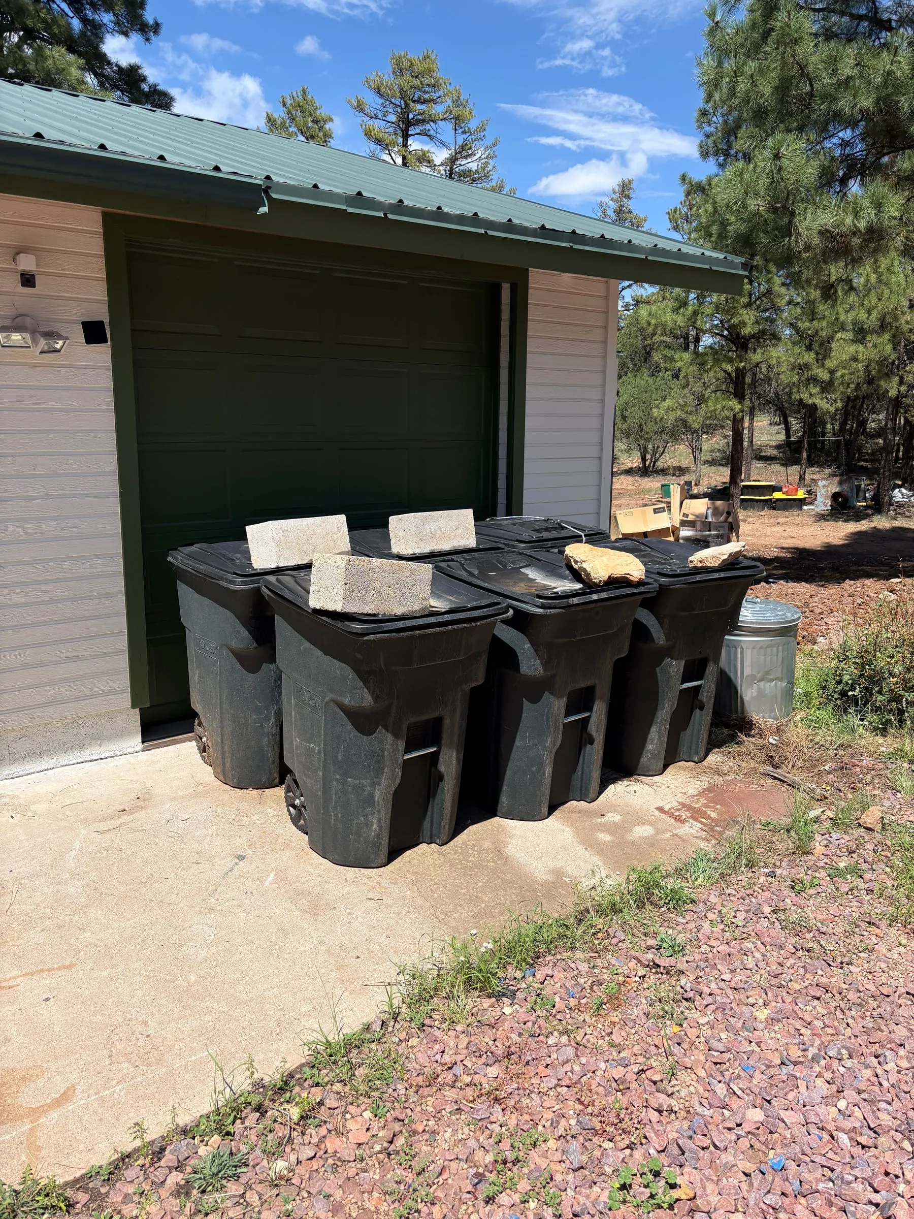 Four black wheeled bins and a metal can standing in a row against the garage wall on the gravel, each lid weighted shut with a rock or a paving block.